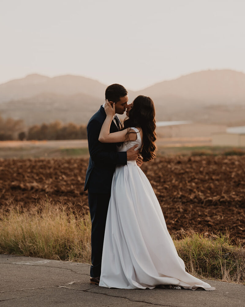 Bride and groom kiss in front of a field and barn during golden hour portraits at their wedding in ramona ca
