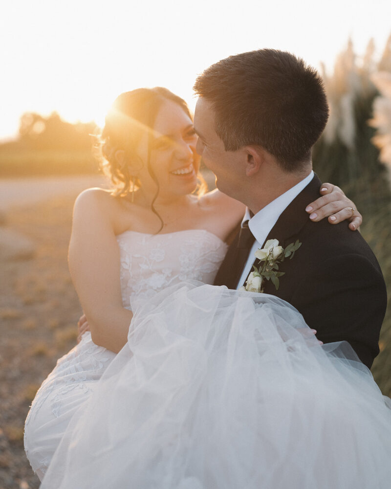 groom carries his bride and smiles lovingly at her during sunset portraits at their wedding in ramona