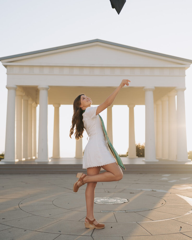 PLNU Grad photos College Senior Graduate throwing her cap in the air while standing in front of the Point Loma Nazarene Greek Amphitheater