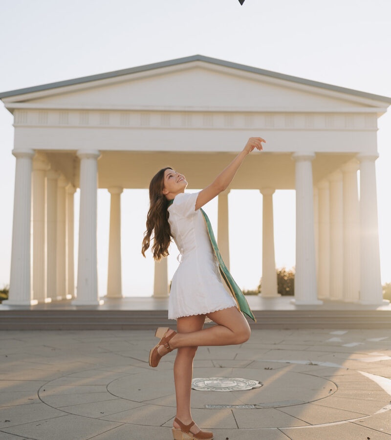 PLNU Grad photos College Senior Graduate throwing her cap in the air while standing in front of the Point Loma Nazarene Greek Amphitheater