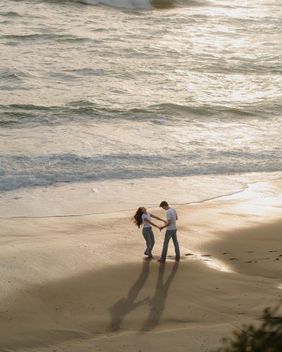 engaged couple dances on the beach in a wide photo during their playful cinematic engagement session at sunset