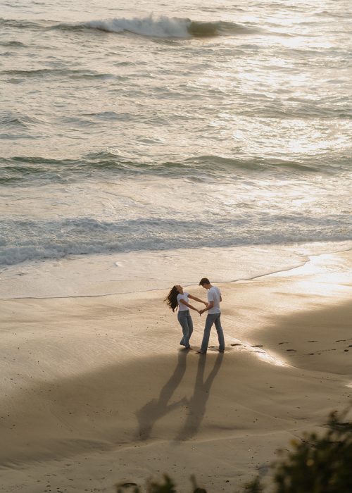 engaged couple dances on the beach in a wide photo during their playful cinematic engagement session at sunset