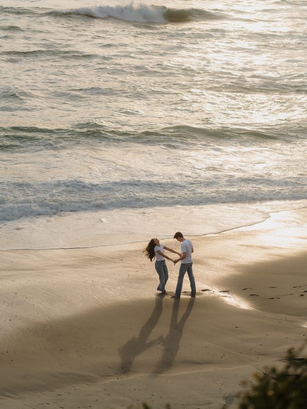 engaged couple dances on the beach in a wide photo during their playful cinematic engagement session at sunset