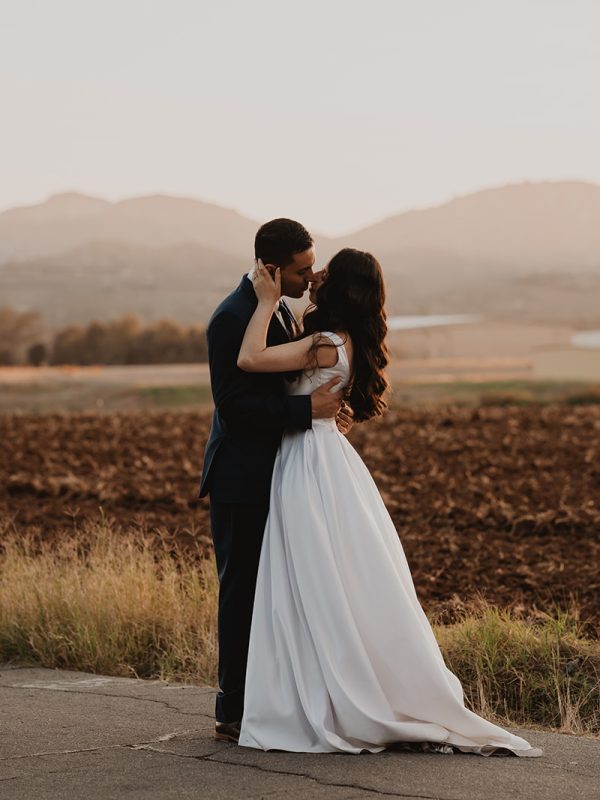 Bride and groom kiss in front of a field and barn during golden hour portraits at their wedding in ramona ca