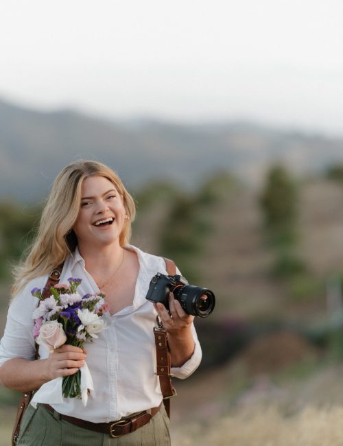 Photographer Alaina Yard of Alaina Yard photography smiles at the camera while holding wedding bouquet and camera in a winery